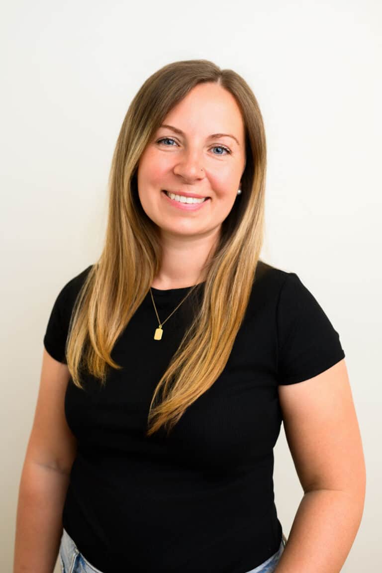 Woman with long, straight light brown hair, wearing a black t-shirt, gold necklace, and smiling in front of a plain light background.